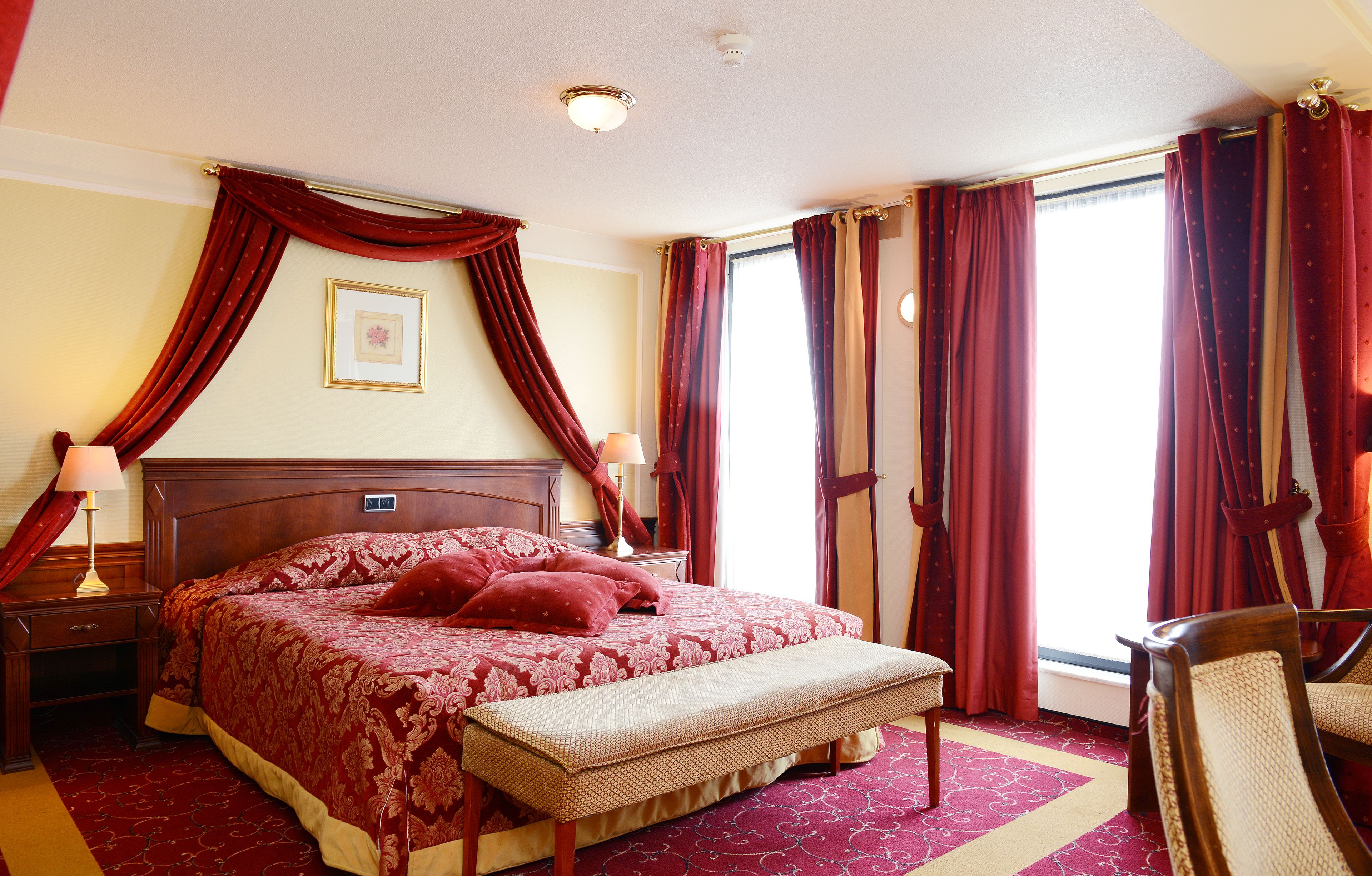 Ein Hotelzimmer mit einem großen Bett, einer Bank, einem Nachttisch, einer Lampe und einem Fenster mit Vorhängen.