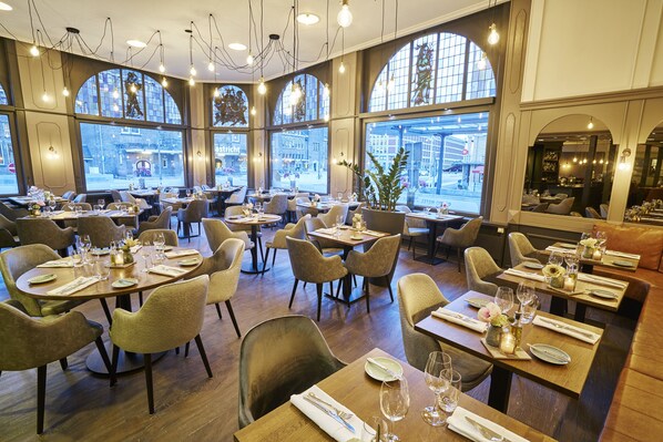A restaurant with multiple dining tables set for guests, large windows offering a view of the street, and modern lighting fixtures.