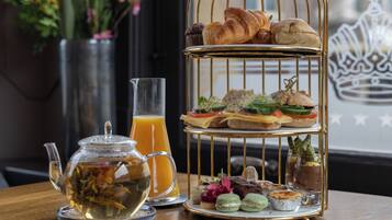 A tiered dessert stand with pastries and sandwiches, a glass teapot with tea, and a glass of orange juice on a wooden table.