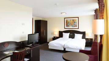 Een hotelkamer met een bed, een bureau met een computer, een stoel, een nachtkastje met een lamp en een schilderij aan de muur.