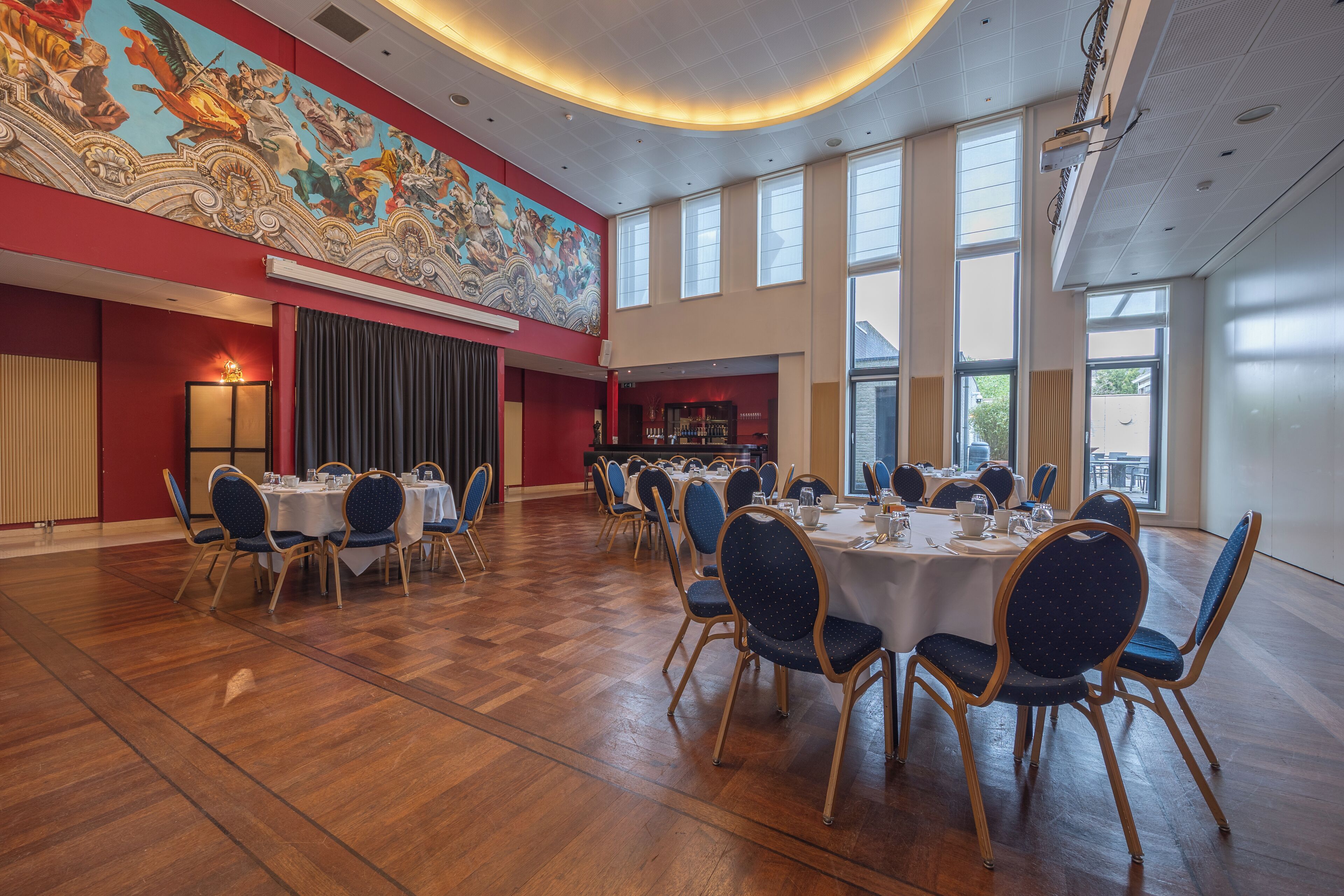 A spacious hall with round tables and chairs, a large mural on the wall, and large windows allowing natural light.