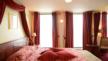 Een hotelkamer met een groot bed, een stoel, een lamp en uitzicht op het omliggende landschap door de ramen.