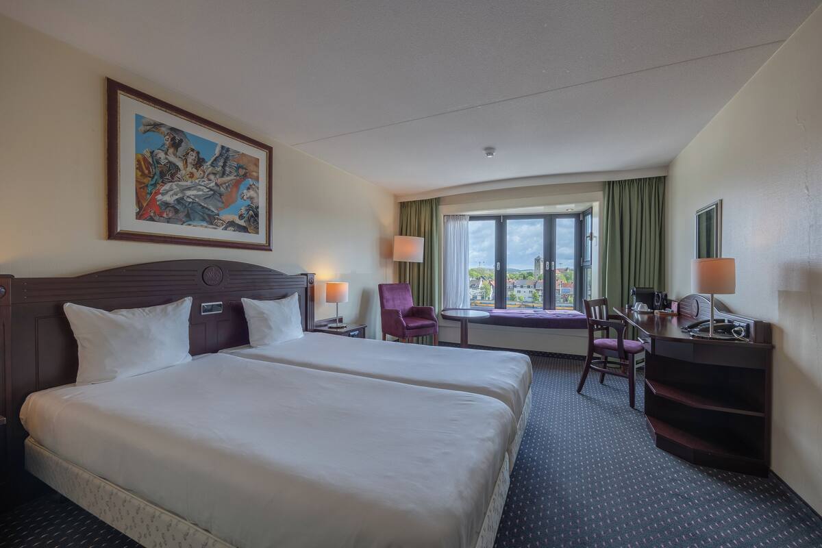 A hotel room with two beds, a desk, a chair, a lamp, a painting on the wall, and a window with a view.