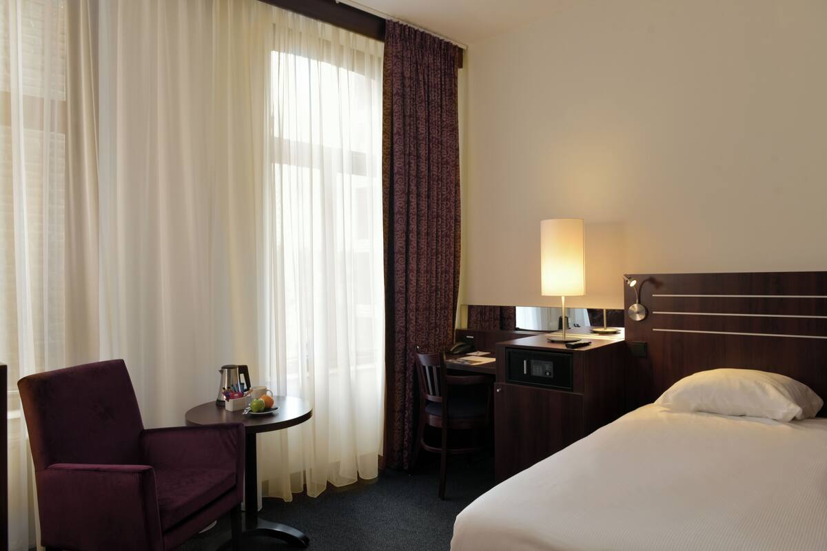 A hotel room with a bed, a desk, a chair, a lamp, and a small table with a fruit bowl.