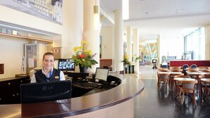 A hotel reception area with a curved desk, multiple monitors, a computer, and a plant. There are several tables and chairs in the background. - Amrâth Grand Hotel de l'Empereur (Maastricht)