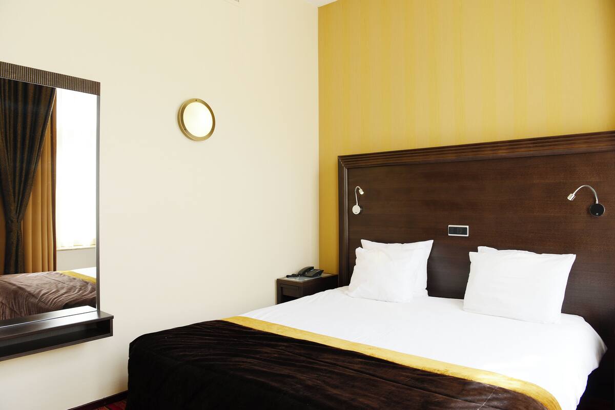 A hotel room with a large bed, a bedside table with a phone, a mirror, and a wall-mounted light fixture.