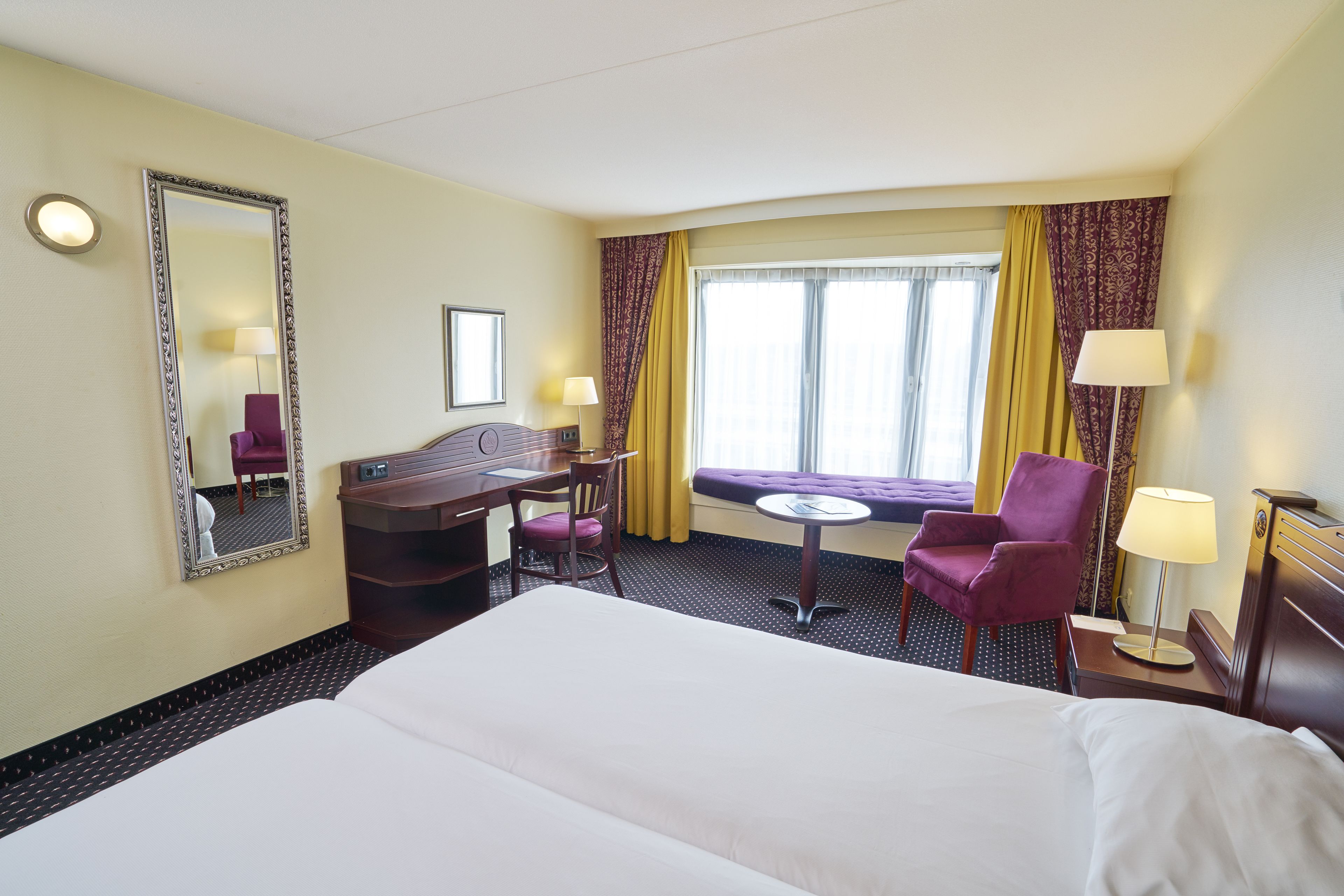 A hotel room with a large bed, a desk, two chairs, a small table, and a window with curtains.