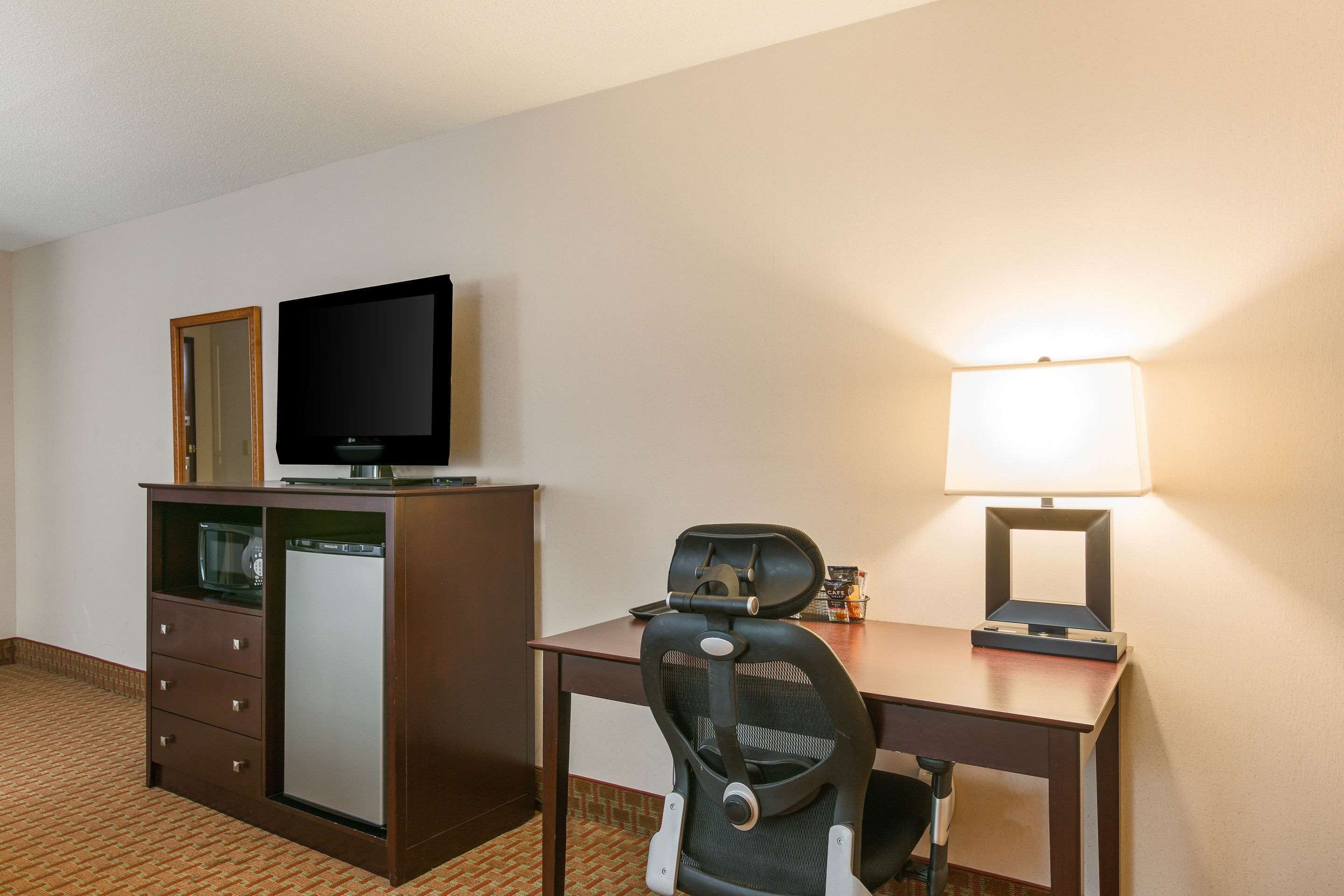 A hotel room with a desk, a chair, a television on a cabinet, a lamp, and a refrigerator.