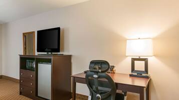 A hotel room with a desk, a chair, a television on a cabinet, a lamp, and a refrigerator.