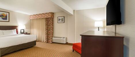 A hotel room with a bed, a desk, a chair, a television, a lamp, and an air conditioning unit.