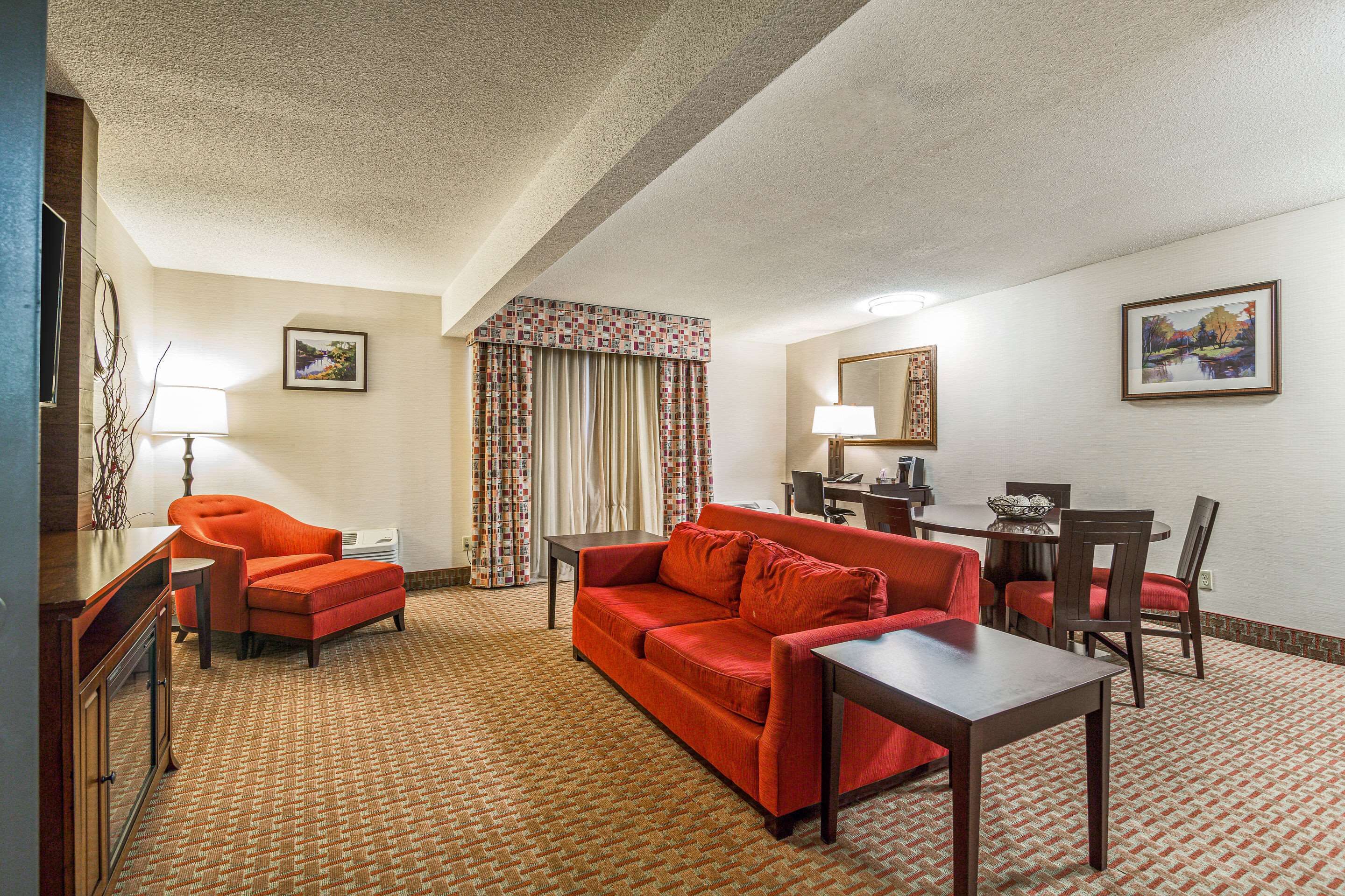 A hotel room with a red sofa, a wooden coffee table, a desk with a lamp, a dining table with chairs, and a patterned carpet.