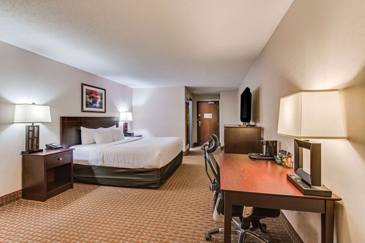 A hotel room with a bed, two bedside lamps, a desk with a chair, and a door leading to another room.