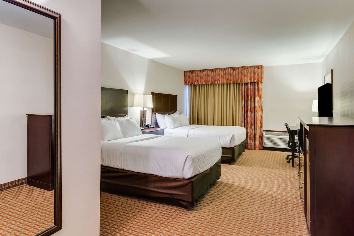 A hotel room with two beds, a desk, a chair, a mirror, and a window with curtains.