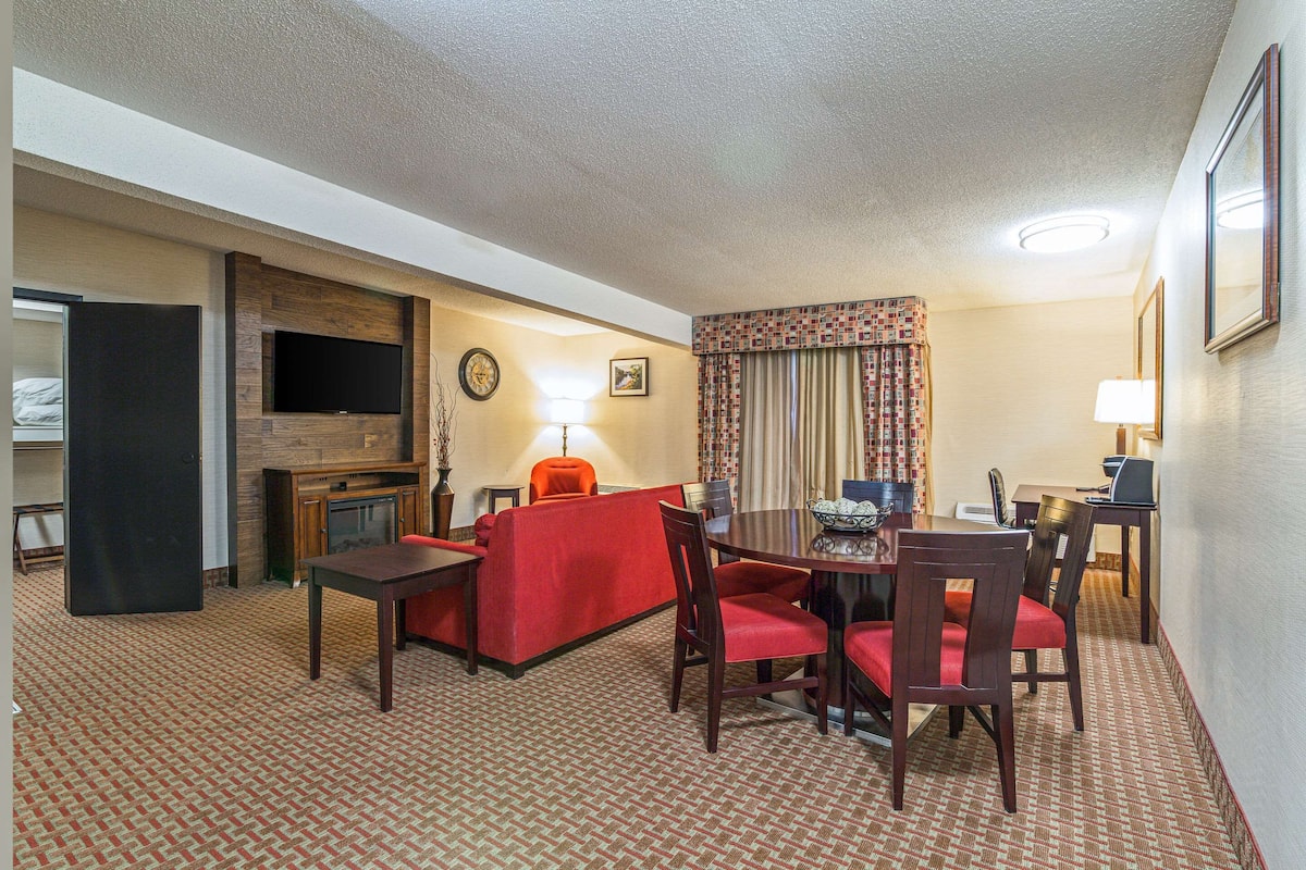 A hotel room with a dining area, a TV mounted on the wall, a fireplace, and a desk with a lamp.
