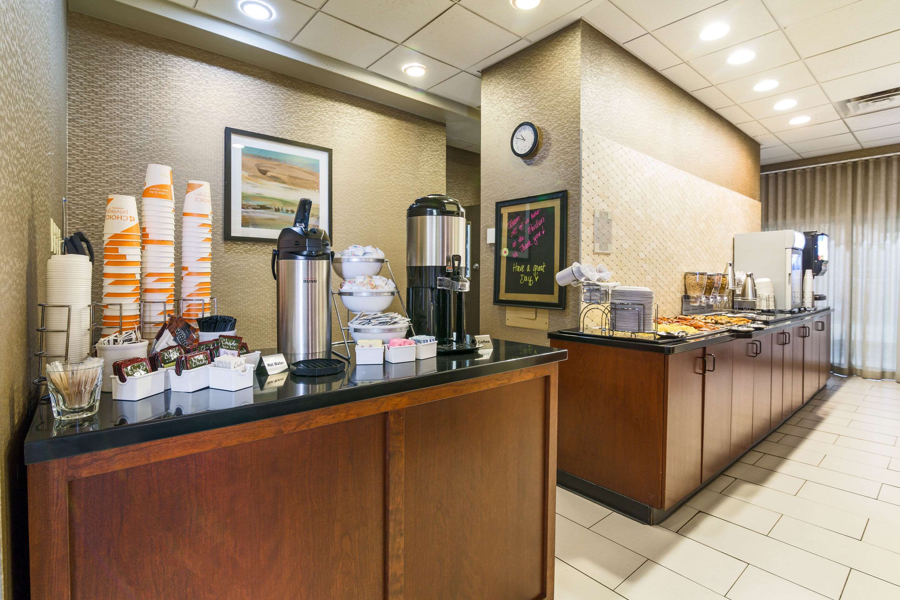 Espaço de café da manhã de hotel com serviço de buffet, incluindo máquinas de café, um relógio e uma lousa com uma mensagem.