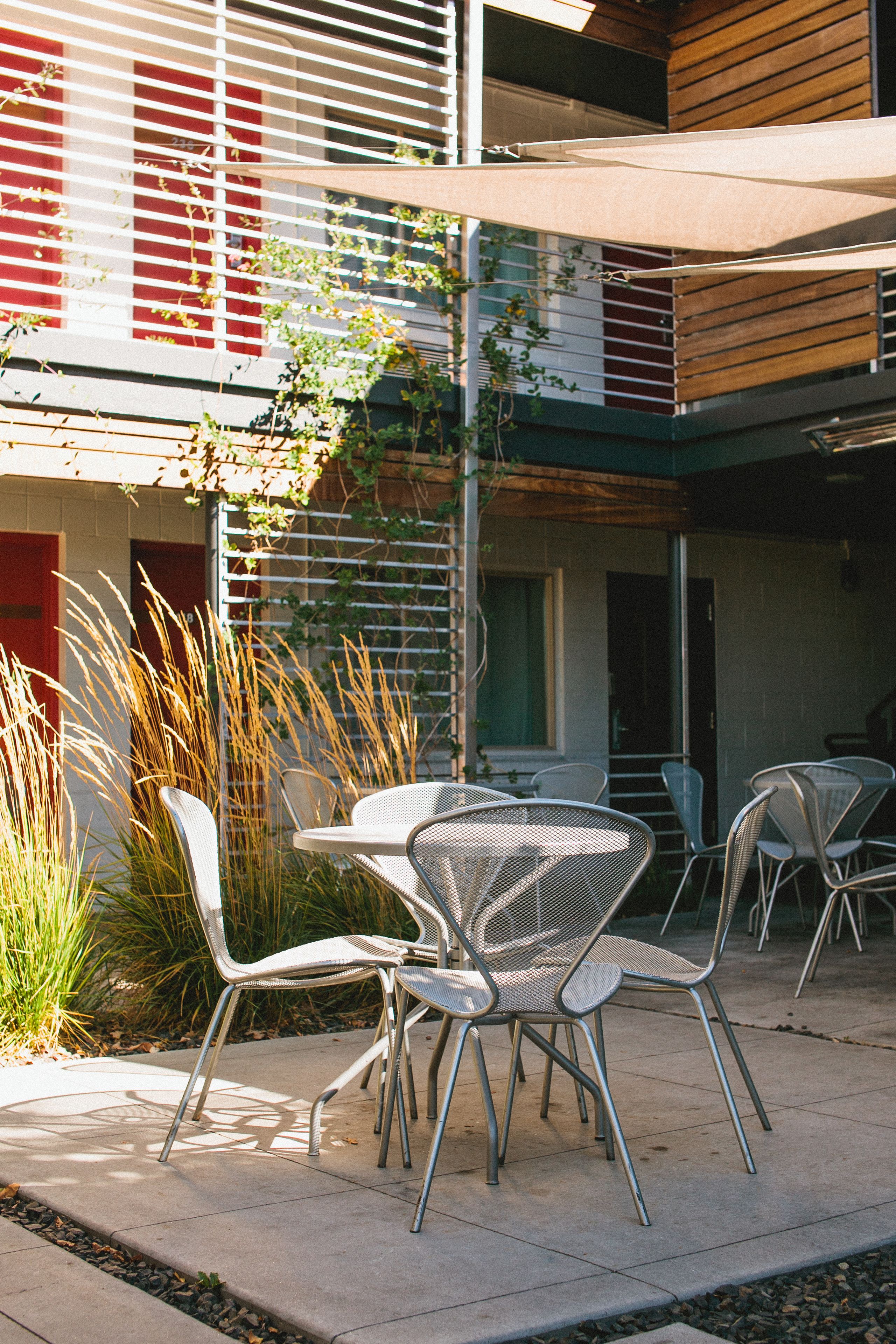 An outdoor seating area with metal chairs and a table, surrounded by plants and a building with red and white walls.