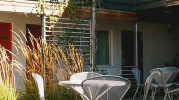 An outdoor seating area with metal chairs and a table, surrounded by plants and a building with red and white walls.