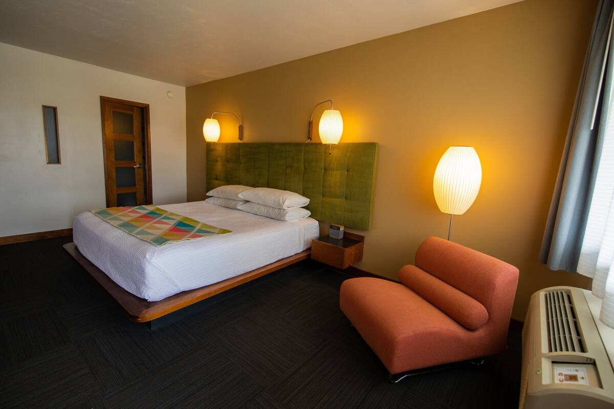 A hotel room with a bed, a nightstand, a chair, and a wall-mounted lamp.