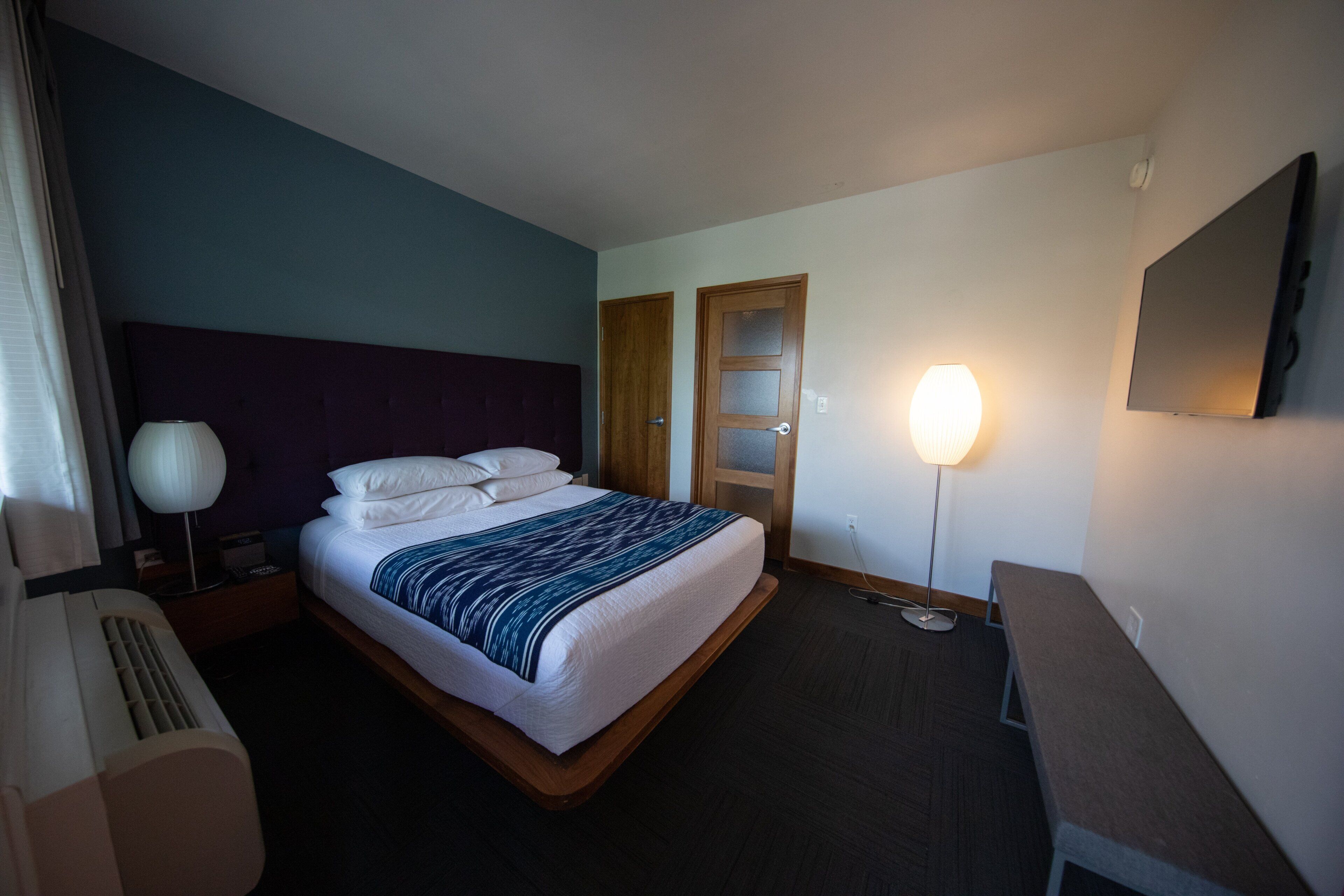 Habitació d'hotel amb llit, taulell de nit, làmpada i televisió de pantalla plana penjada a la paret.