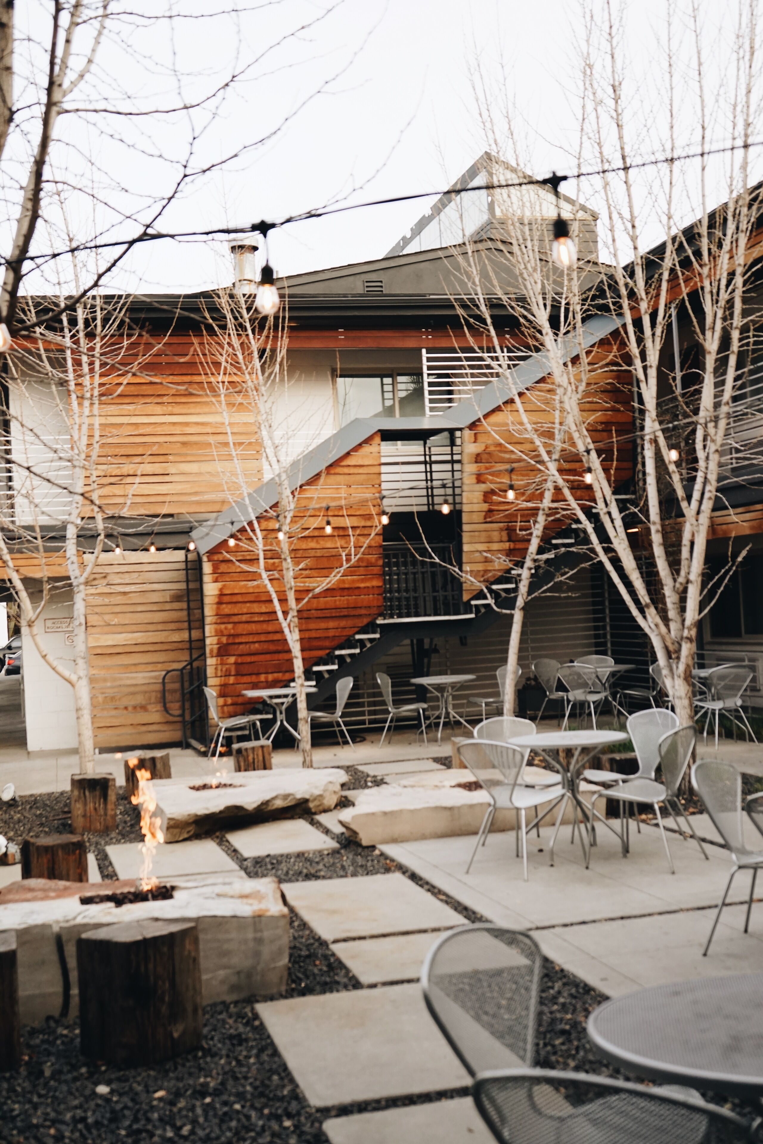 An outdoor patio area with stone flooring, metal chairs, a fire feature, and a wooden building with a staircase.