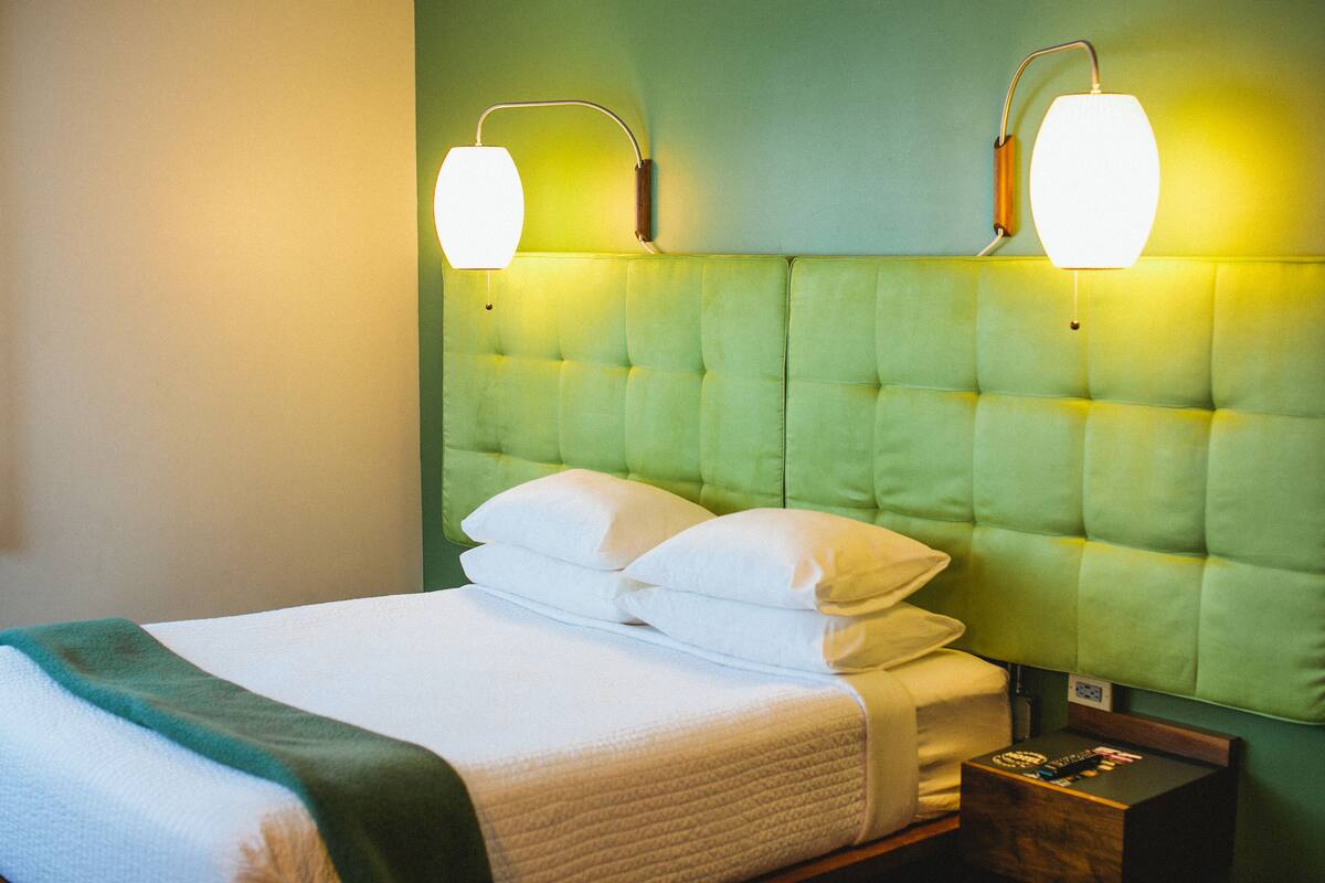 A neatly made bed with white linens and pillows, a green upholstered headboard, and two wall-mounted lamps.