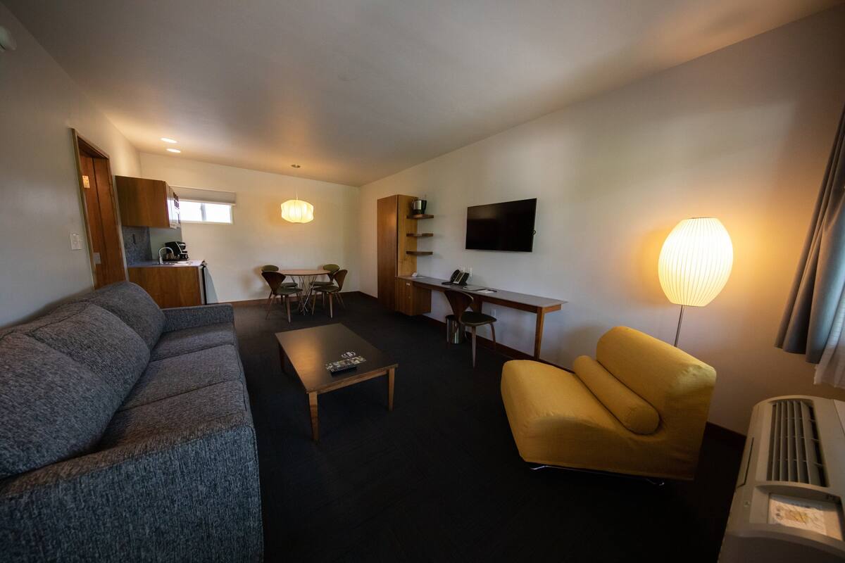 A compact living space with a sofa, a yellow armchair, a dining table with chairs, a TV mounted on the wall, and a kitchenette area.