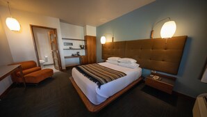 Habitación de hotel con una cama grande, un escritorio, una silla, una mesita de noche y una mesilla auxiliar.