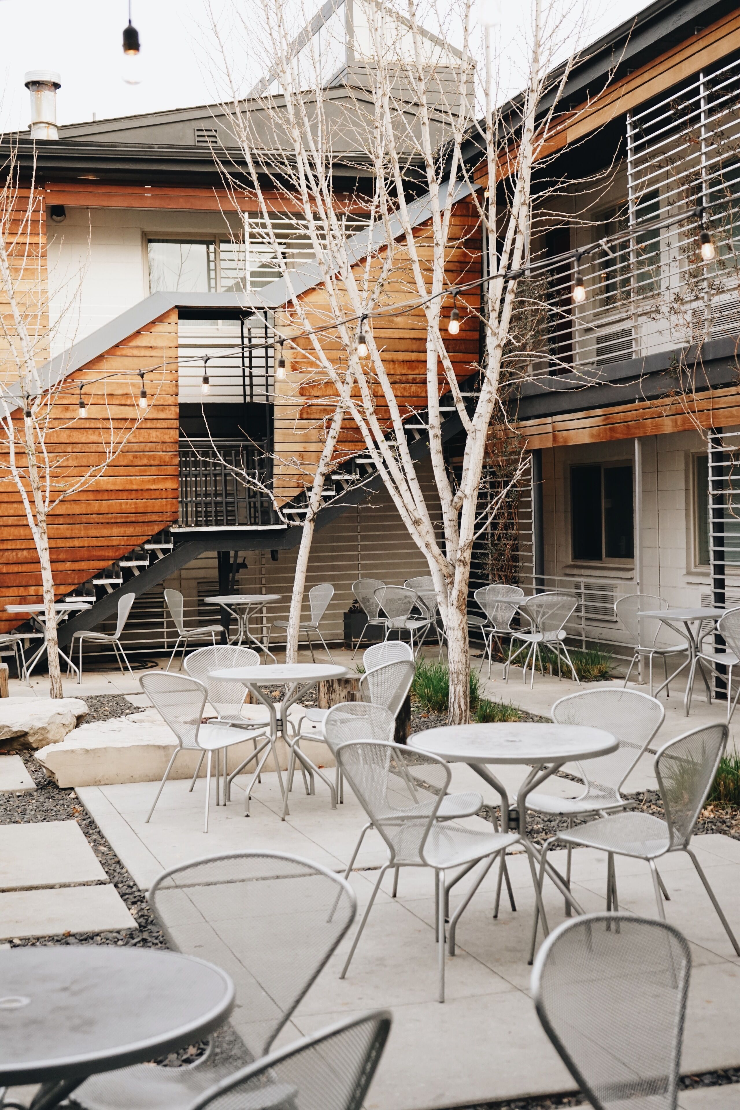 Un patio al aire libre con varias mesas y sillas, una escalera y un edificio con revestimiento de madera y barandillas de metal.