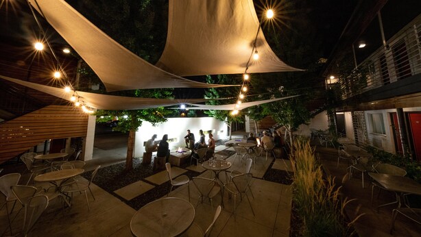 Un área de comedor al aire libre con mesas y sillas, cubierta por grandes toldos blancos, y un edificio al fondo.