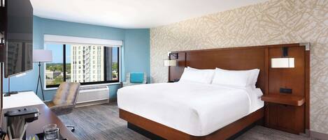A hotel room with a large bed, a desk with a computer, and a view of a cityscape through the window.
