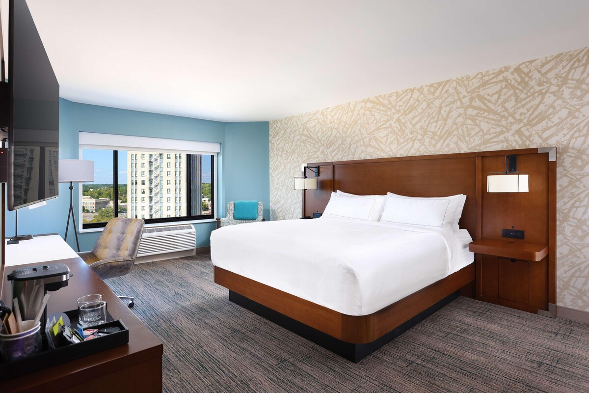 Hotelkamer met een groot bed, een bureau met een computer, een stoel en uitzicht op een stadsbeeld door het raam.