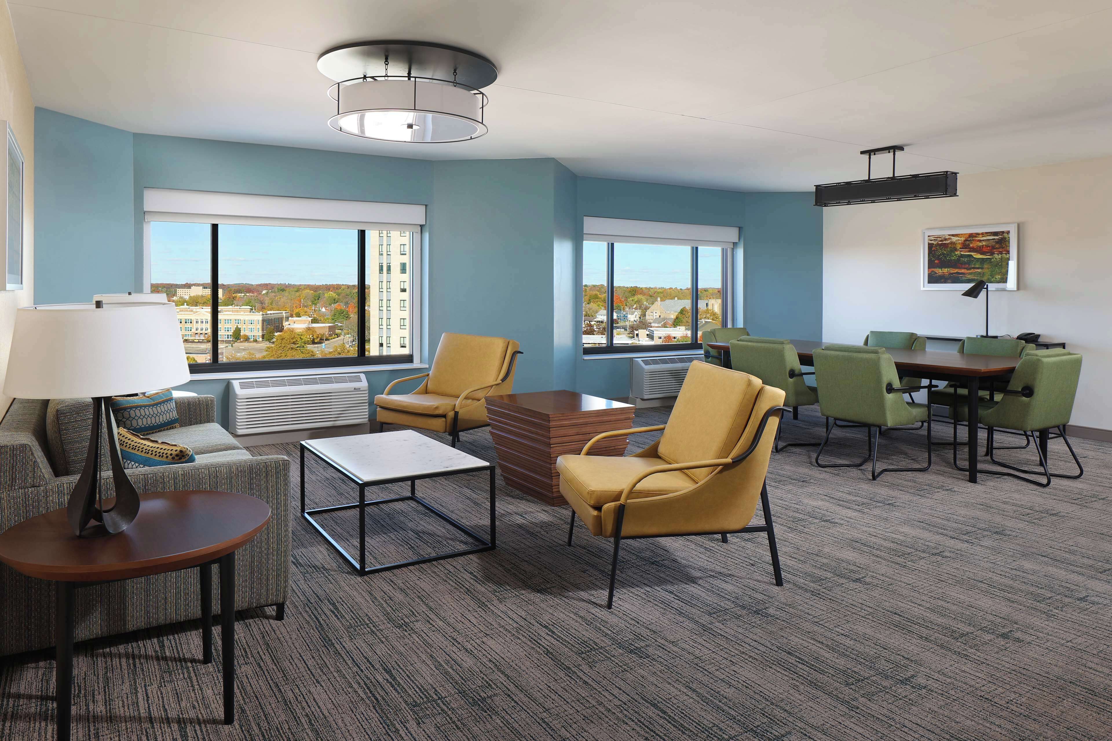 A modern hotel room with a large window, a sofa, a coffee table, and a dining area with chairs.