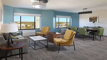 A modern hotel room with a large window, a sofa, a coffee table, and a dining area with chairs.