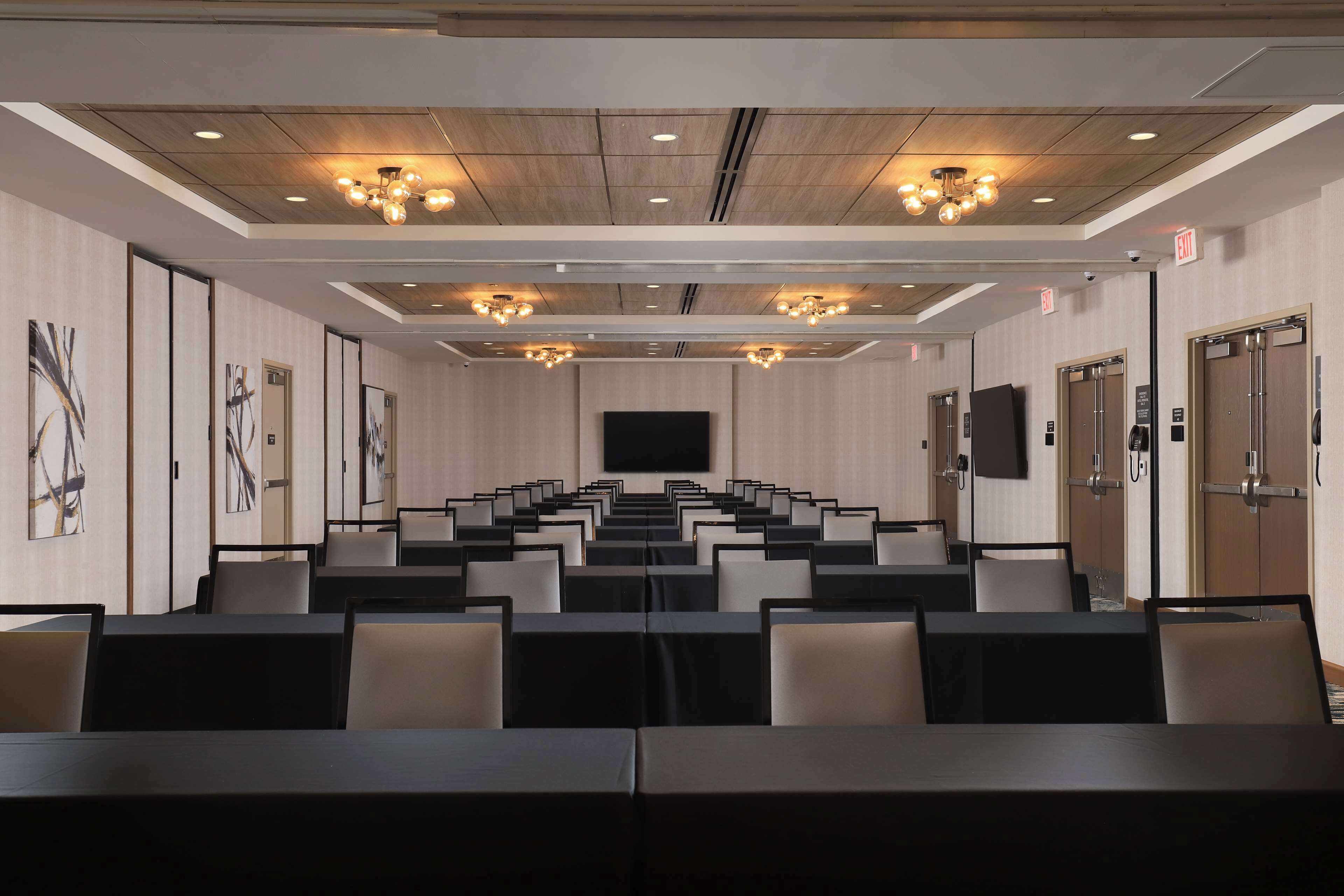 Une salle de conférence avec des rangées de tables et de chaises, un grand écran à l’avant et des luminaires au plafond.