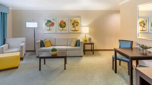 Una sala de estar con sofá, sillón, mesa de centro y mesa de comedor. Hay tres cuadros en la pared, una lámpara y una pequeña mesa con una planta.
