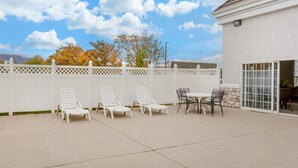 A patio with white lounge chairs and a table, a white fence, and a building with a tiled roof.