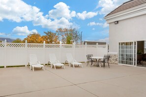 A patio with white lounge chairs and a table, a white fence, and a building with a tiled roof.