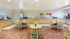 A hotel dining area with multiple tables and chairs, a kitchen counter with appliances, and a patterned carpet.