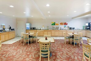 A hotel dining area with multiple tables and chairs, a kitchen counter with appliances, and a patterned carpet.