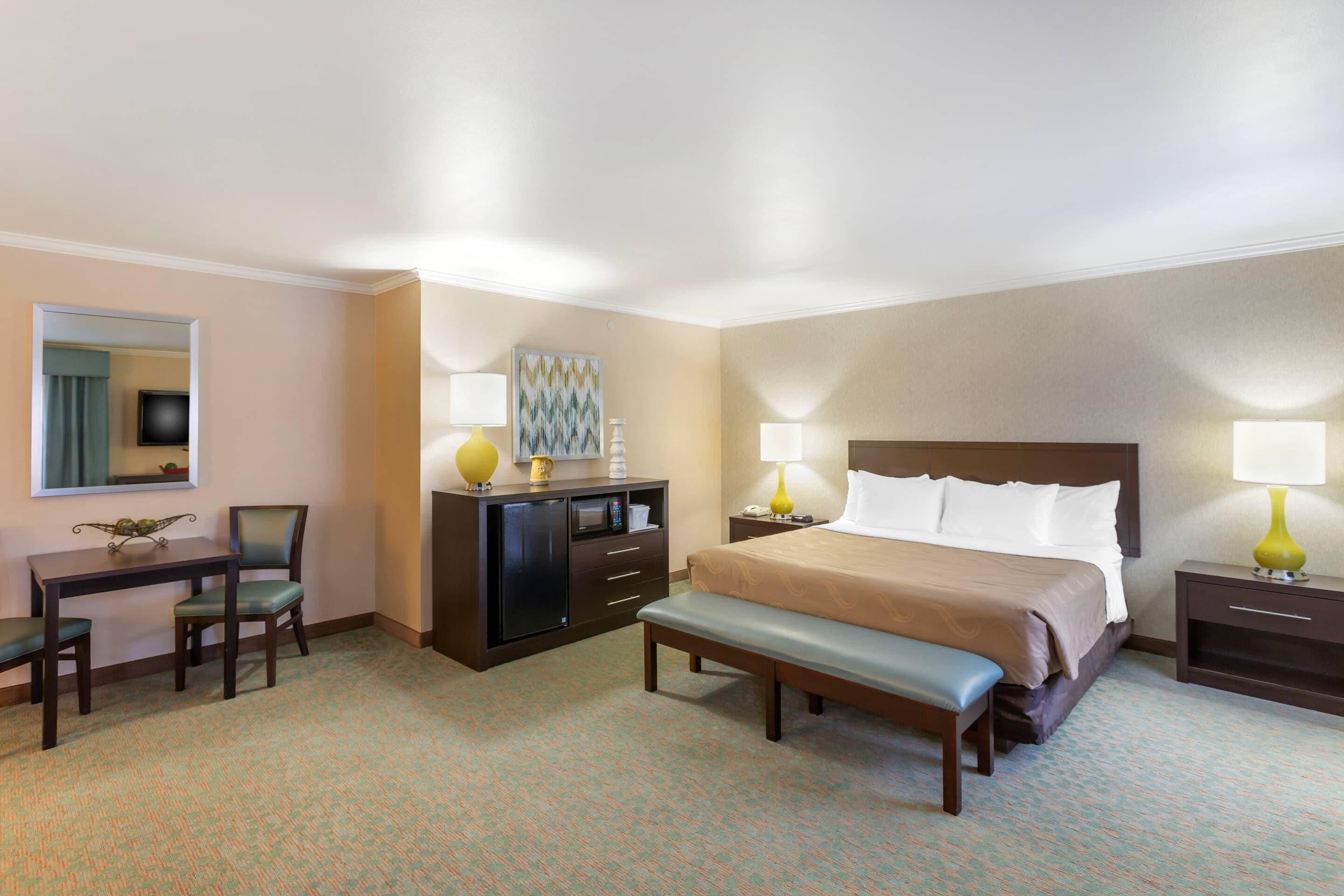 A hotel room with a bed, desk, chair, and a small table with a lamp.