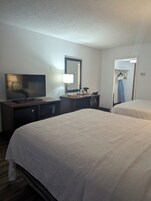 Quarto de hotel com duas camas, uma mesa com televisão, um espelho e uma porta para uma casa de banho com porta-toalhas.