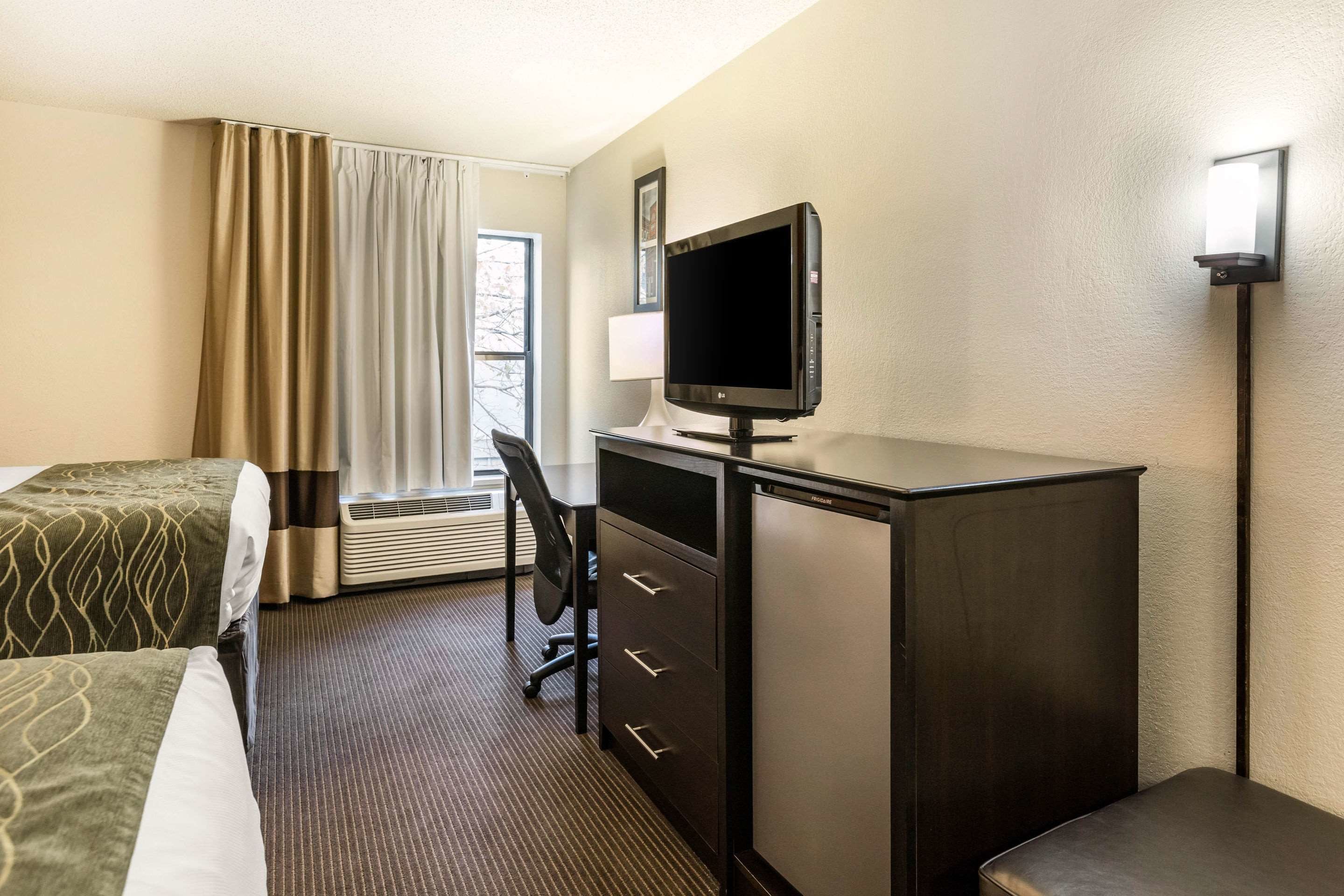 A hotel room with a bed, a desk with a computer, a chair, a lamp, and a window with curtains.