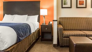 A hotel room with a bed, a nightstand with a lamp, a patterned sofa, and two framed pictures on the wall. - Comfort Inn Wings Stadium (Kalamazoo)