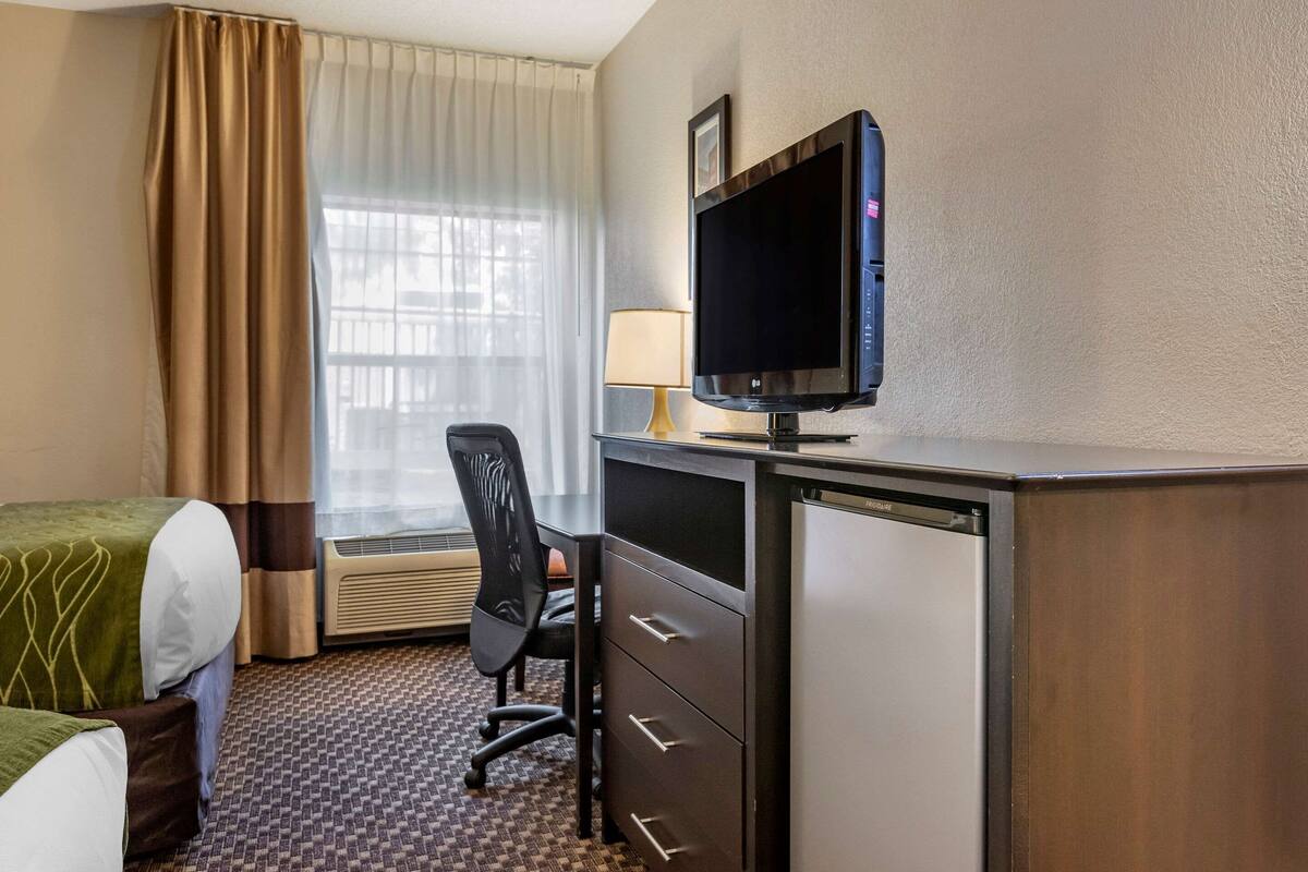 A hotel room with a bed, a desk with a computer and a TV, a chair, a lamp, and a window with curtains.