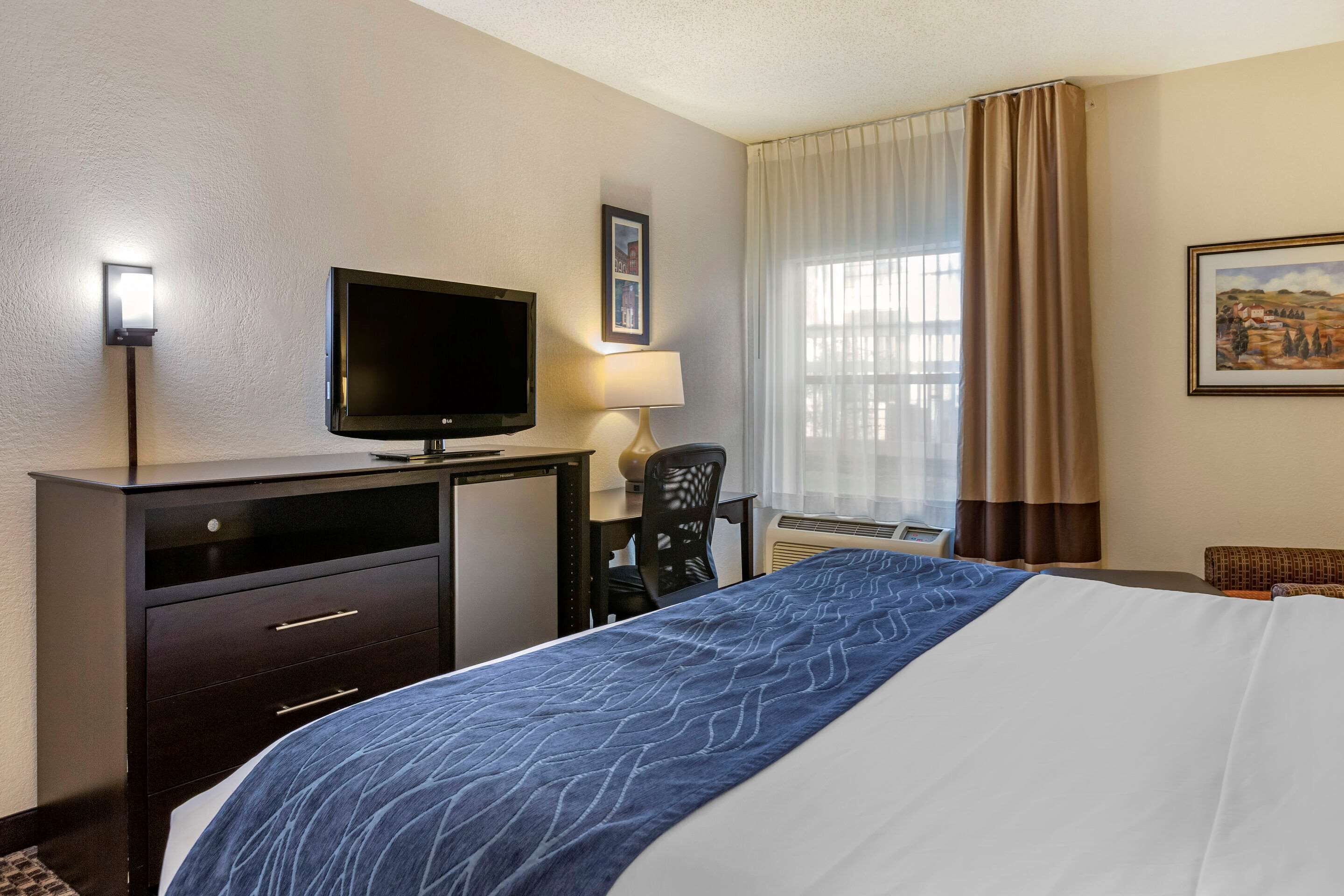Ein Hotelzimmer mit einem Bett, einem Schreibtisch mit Fernseher, einem Sessel und einem Fenster mit Vorhängen.