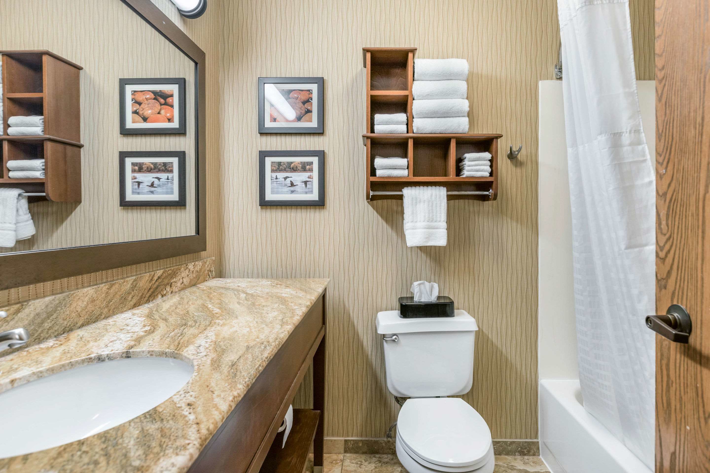 A bathroom with a marble sink, wooden shelves with towels, framed pictures on the wall, and a shower curtain.