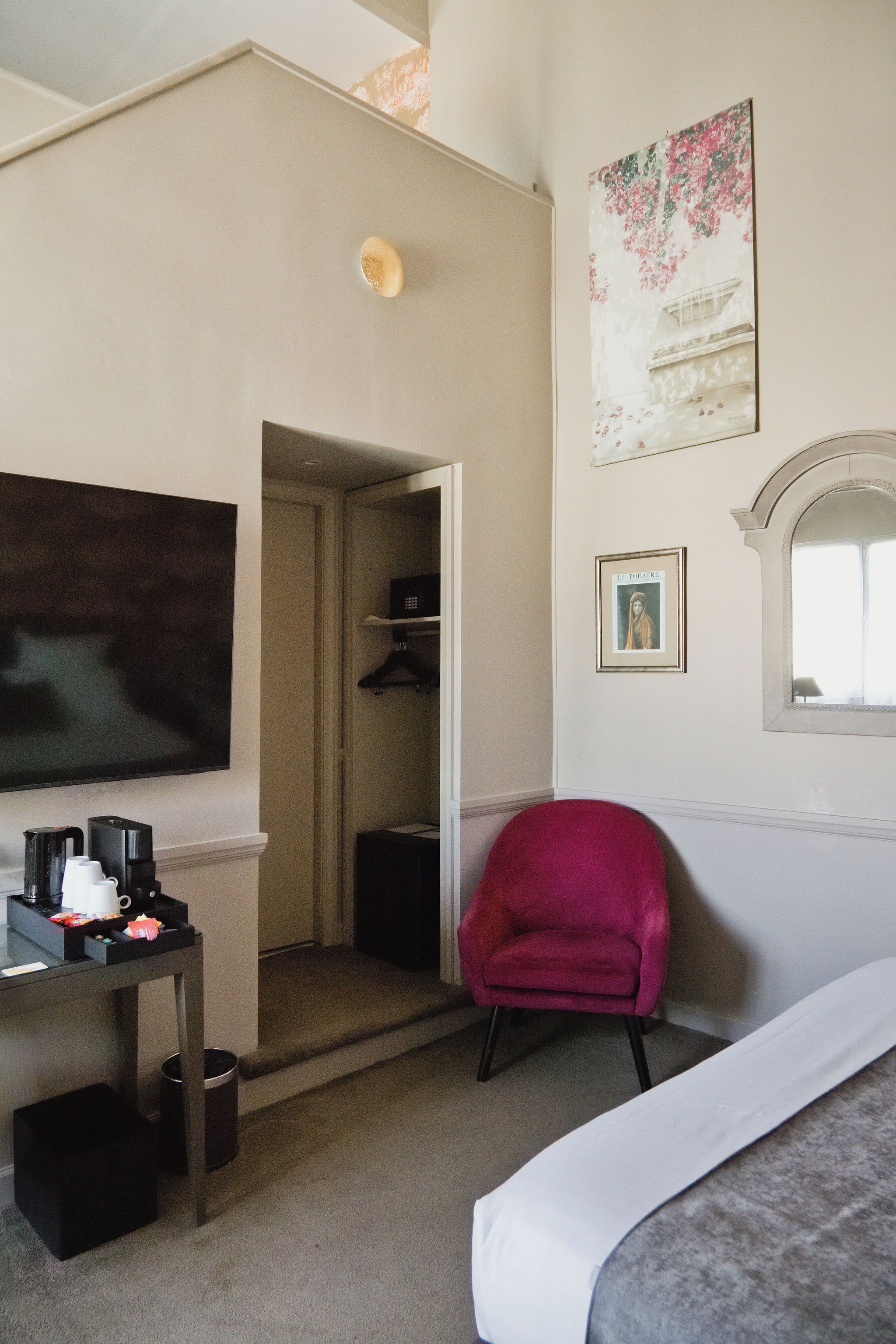Habitación de hotel con cama, escritorio con cafetera, una silla roja, televisor y puerta que da a un armario.