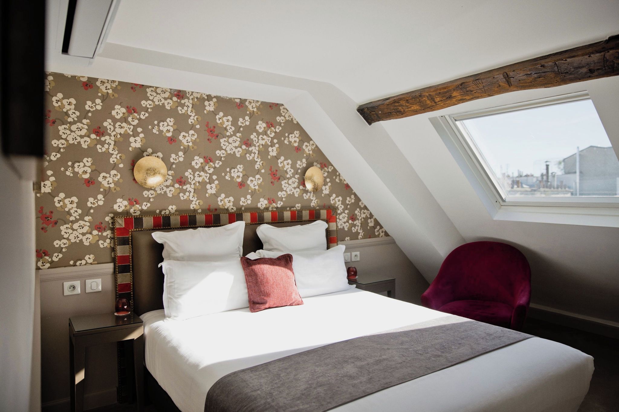 A hotel room with a bed, a red chair, a window, and a floral wallpaper.
