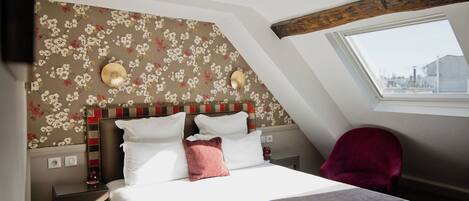 A hotel room with a bed, a red chair, a window, and a floral wallpaper.