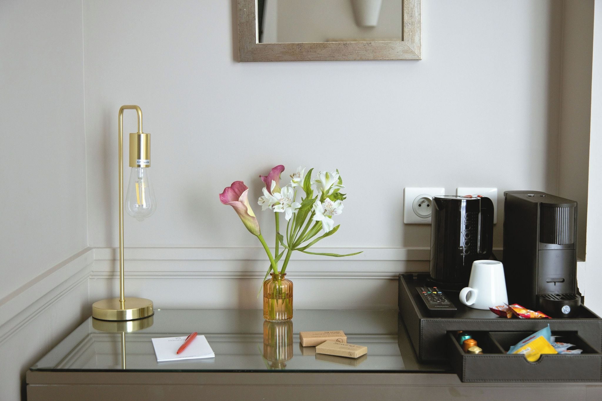 A hotel room with a bedside table featuring a lamp, a vase with flowers, a coffee maker, a remote control, and a cup.
